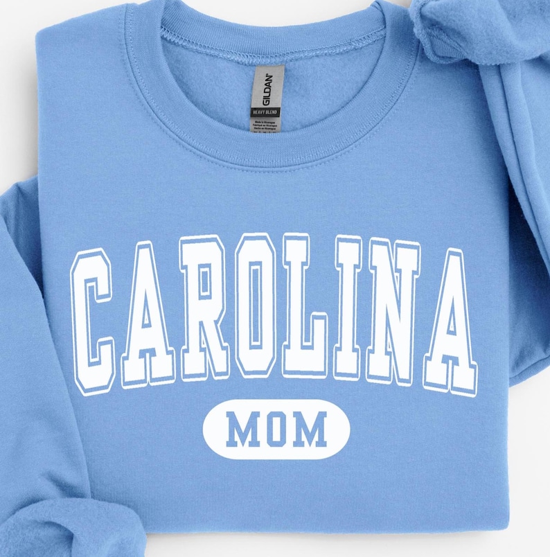 Puede incluir: Una sudadera azul claro con la palabra "CAROLINA" en letras blancas grandes y la palabra "MOM" en un &oacute;valo blanco debajo. La sudadera es de mezcla pesada y tiene cuello redondo.
