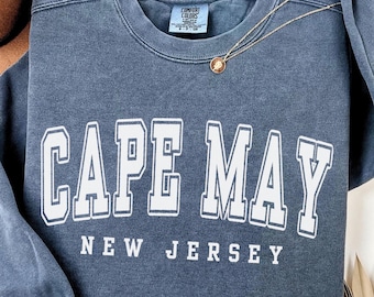 May include: A blue crewneck sweatshirt with a vintage-washed finish. The front features the white text "CAPE MAY" in a large collegiate font, with "NEW JERSEY" printed below in a smaller font. A thin gold chain necklace rests on the neckline.