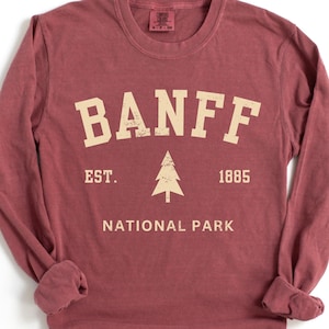 May include: A long-sleeve crew neck t-shirt in a muted brick red color. The front features cream-colored distressed text that reads BANFF, EST. 1885, and NATIONAL PARK, with a central graphic of a pine tree between the year and the abbreviation.