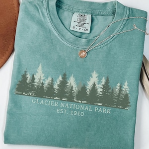 Könnte beinhalten: Ein blaugrünes Kurzarm-T-Shirt mit einer dunkelgrünen Grafik einer Waldsilhouette und dem weißen Text "GLACIER NATIONAL PARK EST. 1910". Das T-Shirt hat einen Rundhalsausschnitt und ein aufgedrucktes Comfort Colors-Etikett am Innenkragen.