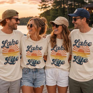 May include: Four people wearing cream-colored t-shirts with a retro sunset graphic and the words "Lake Life" in navy blue. They are standing near a lake at sunset, wearing sunglasses and hats.