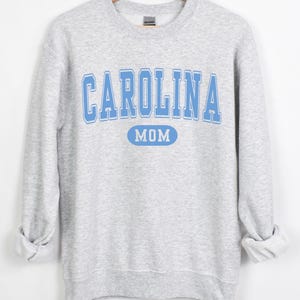 Puede incluir: Sudadera gris claro con las palabras "CAROLINA MOM" en letras de bloque azul claro. La palabra "MOM" est&aacute; en un &oacute;valo azul claro. La sudadera tiene mangas largas y cuello redondo.