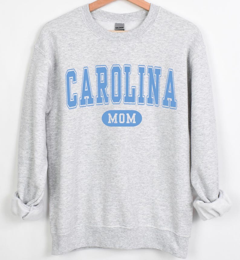Puede incluir: Sudadera gris claro con las palabras "CAROLINA MOM" en letras de bloque azul claro. La palabra "MOM" est&aacute; en un &oacute;valo azul claro. La sudadera tiene mangas largas y cuello redondo.