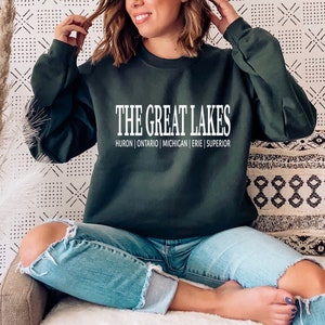 May include: A dark forest green long-sleeve crewneck sweatshirt featuring the white text THE GREAT LAKES followed by the names HURON | ONTARIO | MICHIGAN | ERIE | SUPERIOR printed in a clean, sans-serif font across the center of the chest.
