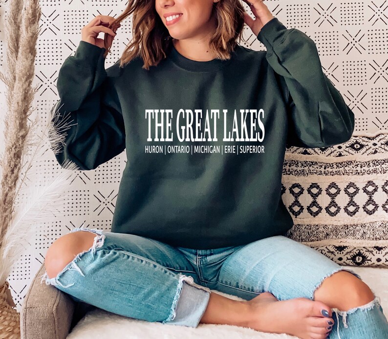 May include: A dark forest green long-sleeve crewneck sweatshirt featuring the white text THE GREAT LAKES followed by the names HURON | ONTARIO | MICHIGAN | ERIE | SUPERIOR printed in a clean, sans-serif font across the center of the chest.