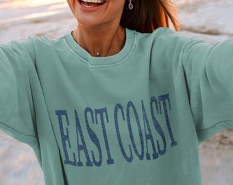 May include: A sage green crewneck sweatshirt featuring the text EAST COAST printed in a dark blue, distressed font across the chest. The garment has long sleeves, a ribbed collar, and a relaxed fit, showcasing a solid colour throughout the fabric.