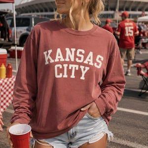 May include: A maroon long-sleeve sweatshirt with the words "KANSAS CITY" in white block letters. The person is wearing denim shorts and holding a red cup. The background includes a tailgate setup with food and drinks.