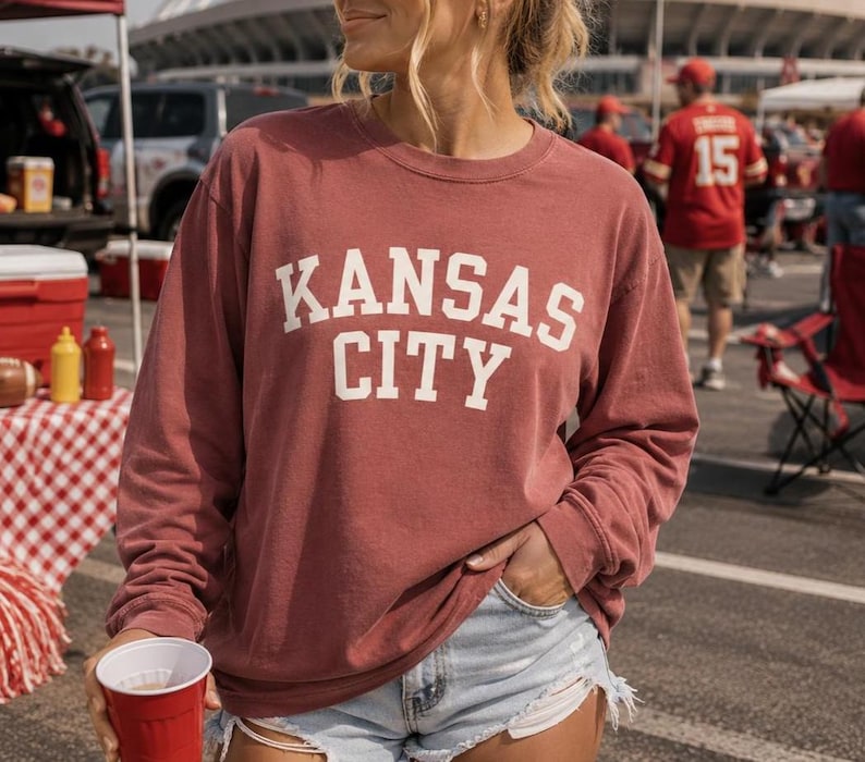 May include: A maroon long-sleeve sweatshirt with the words "KANSAS CITY" in white block letters. The person is wearing denim shorts and holding a red cup. The background includes a tailgate setup with food and drinks.