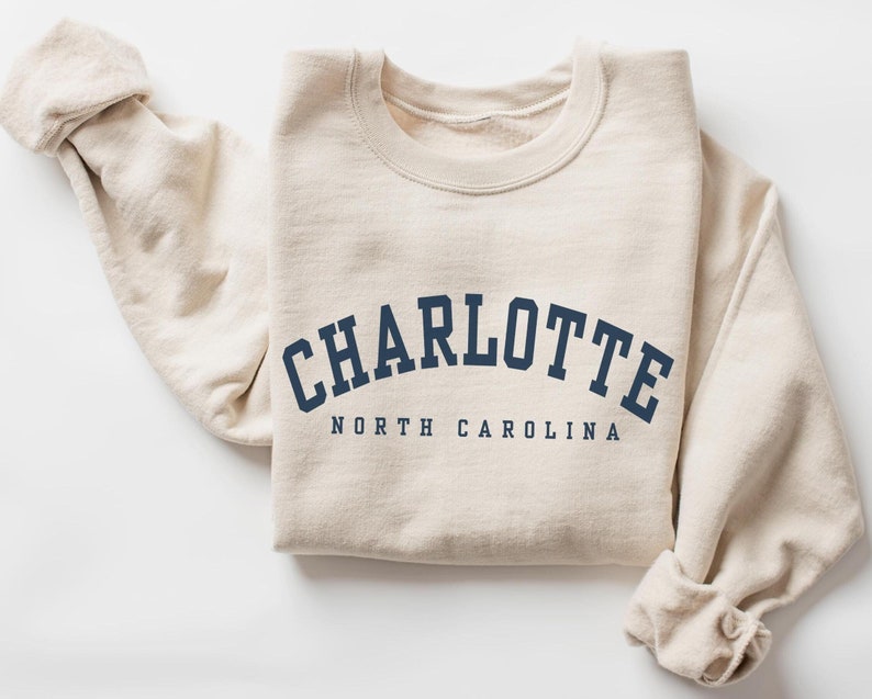 May include: A cream-colored sweatshirt with the word "CHARLOTTE" in navy blue arched across the chest, with "NORTH CAROLINA" printed below. The sweatshirt has long sleeves and a crew neck.