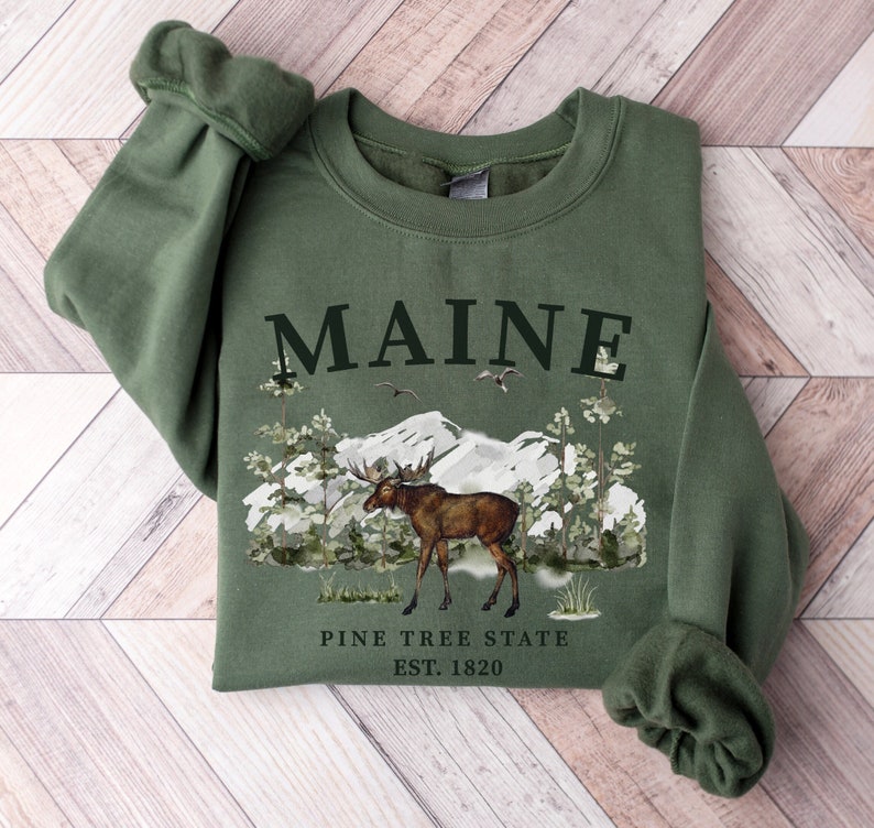 May include: A green sweatshirt with a moose and mountain scene. The text "MAINE PINE TREE STATE EST. 1820" is printed on the front.