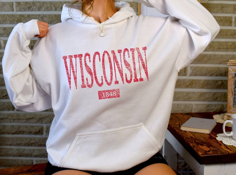 Puede incluir: Una sudadera con capucha blanca que presenta la palabra WISCONSIN impresa en una fuente roja desgastada en el pecho. Debajo del texto hay un recuadro rojo con el n&uacute;mero 1848 en blanco. La prenda incluye un bolsillo tipo canguro frontal.