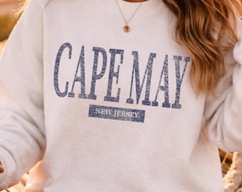 May include: A white crewneck sweatshirt featuring the text CAPE MAY in a distressed blue font, with the words NEW JERSEY printed in a smaller blue rectangular box directly underneath the main text, creating a casual and simple aesthetic.
