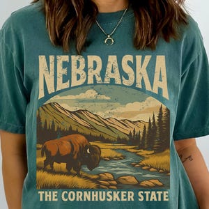 Op de afbeelding: Tealkleurig T-shirt met een afbeelding van een bizon in een landschap met bergen en een rivier. De tekst "NEBRASKA" staat in een boog boven de scène, met "THE CORNHUSKER STATE" eronder. Het ontwerp heeft een vintage, outdoor esthetiek.