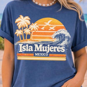 May include: Blue t-shirt featuring a tropical sunset design with palm trees, waves, and the text "Isla Mujeres MEXICO". The graphic is in shades of orange, yellow, and white. The shirt has rolled-up sleeves.