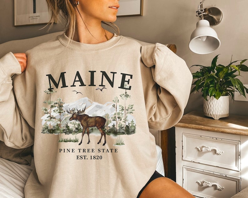 May include: A beige sweatshirt with a graphic print of a moose in a forest setting. The text "MAINE PINE TREE STATE EST. 1820" is printed on the sweatshirt.