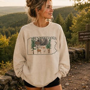 Peut inclure: Un sweat-shirt &agrave; col rond beige pr&eacute;sentant le contour graphique de l'&Eacute;tat de Pennsylvanie. &Agrave; l'int&eacute;rieur du contour, le texte PENNSYLVANIA est imprim&eacute; au-dessus d'une illustration d'un chalet, de sapins et de plusieurs cerfs en for&ecirc;t.