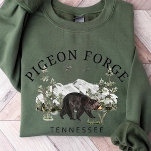 K&ouml;nnte beinhalten: Ein olivgr&uuml;nes Sweatshirt mit Rundhalsausschnitt und schwarzem Grafikdruck. Das Design zeigt den Text PIGEON FORGE in einem Bogen &uuml;ber einer Berglandschaft mit Kiefern, zwei V&ouml;geln und einem Schwarzb&auml;ren. Das Wort TENNESSEE steht darunter.