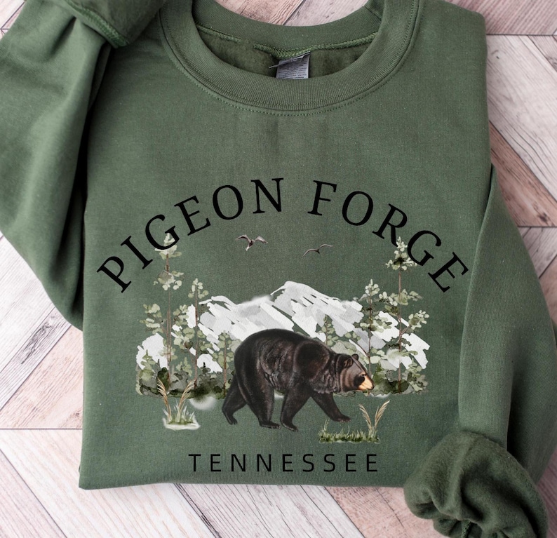 K&ouml;nnte beinhalten: Ein olivgr&uuml;nes Sweatshirt mit Rundhalsausschnitt und schwarzem Grafikdruck. Das Design zeigt den Text PIGEON FORGE in einem Bogen &uuml;ber einer Berglandschaft mit Kiefern, zwei V&ouml;geln und einem Schwarzb&auml;ren. Das Wort TENNESSEE steht darunter.