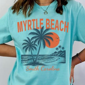 May include: A light blue t-shirt featuring a graphic print with the text MYRTLE BEACH in orange letters at the top and South Carolina in orange script at the bottom. The design includes two palm trees, a large orange sun, and a coastal scene.