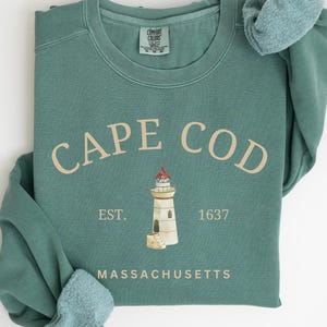 Puede incluir: Una sudadera de color verde azulado con un diseño gráfico blanco de un faro y el texto "CAPE COD EST. 1637 MASSACHUSETTS".