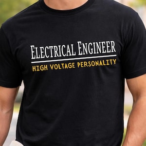 Może przedstawiać: Czarny t-shirt z krótkim rękawem z białym napisem "ELECTRICAL ENGINEER" w górnym wierszu, po którym następuje pozioma biała linia oraz żółty napis "HIGH VOLTAGE PERSONALITY" w dolnym wierszu, wyśrodkowany na klatce piersiowej.