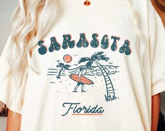 May include: A cream-colored t-shirt featuring a distressed graphic print with the text "SARASOTA" in dark blue and "Florida" in blue. The design depicts a person holding a surfboard on a beach with palm trees, a sun, and ocean waves.