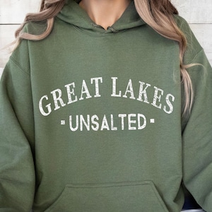 Puede incluir: Una sudadera con capucha de color verde oliva que presenta el texto blanco desgastado "GREAT LAKES" arqueado sobre la palabra "UNSALTED", con un pequeño cuadrado a cada lado de la palabra. La prenda incluye un bolsillo frontal tipo canguro.