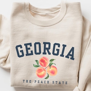 May include: A beige crewneck sweatshirt featuring the dark blue text GEORGIA in an arched font above a watercolor illustration of three peaches with green leaves and the dark blue text THE PEACH STATE printed below the fruit graphic.