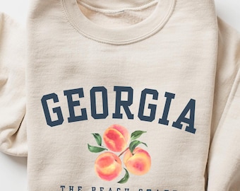 Könnte beinhalten: Ein beigefarbenes Sweatshirt mit Rundhalsausschnitt, das den dunkelblauen Schriftzug GEORGIA in gebogener Schrift über einer Aquarellillustration von drei Pfirsichen mit grünen Blättern und dem dunkelblauen Text THE PEACH STATE zeigt.