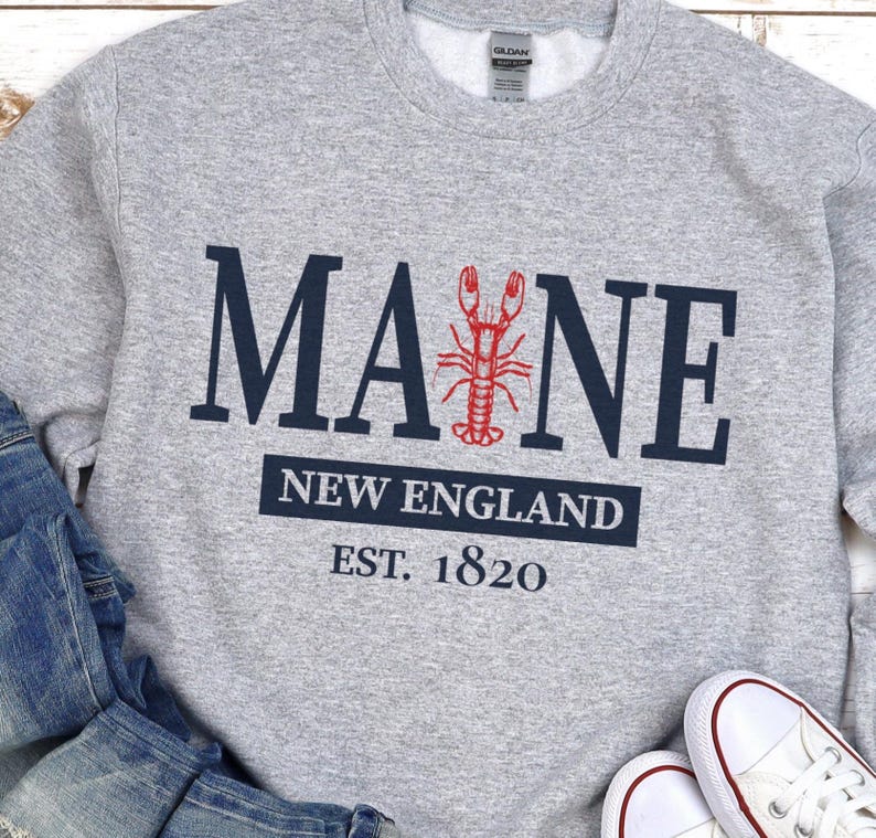 May include: A light grey crewneck sweatshirt featuring the dark blue text MAINE with a red lobster graphic replacing the letter I. Below, a dark blue rectangular block contains the white text NEW ENGLAND and the text EST. 1820 in dark blue.