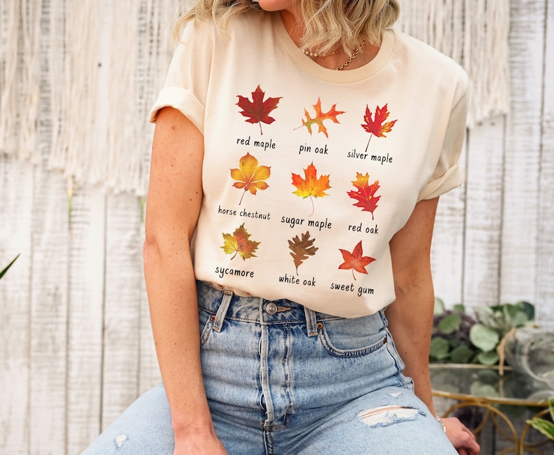 Op de afbeelding: Een beige T-shirt met een afbeelding van verschillende soorten bladeren en hun namen. De bladeren zijn in tinten rood, oranje, geel en bruin. De tekst op het shirt luidt: "red maple", "pin oak", "silver maple", "horse chestnut", "sugar maple", "red oak", "sycamore", "white oak" en "sweet gum".