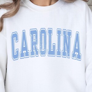 Może przedstawiać: Biała bluza z niebieskim nadrukiem "CAROLINA" w stylu vintage.