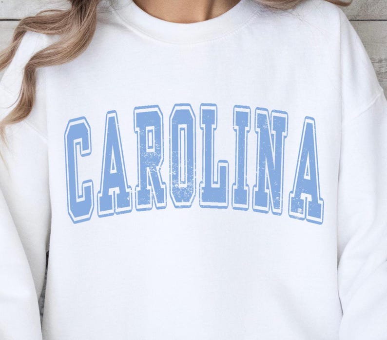 Może przedstawiać: Biała bluza z niebieskim nadrukiem "CAROLINA" w stylu vintage.