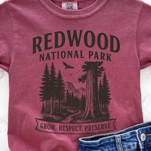 May include: A maroon short-sleeve t-shirt featuring a black graphic of Redwood National Park with trees, mountains, and a bird. The text reads REDWOOD NATIONAL PARK and GROW, RESPECT, PRESERVE. A Comfort Colors brand label is visible at the neck.