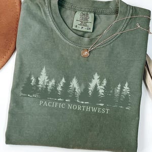 Könnte beinhalten: Ein salbeigrünes T-Shirt mit Rundhalsausschnitt und einem Siebdruck-Motiv einer Waldsilhouette sowie dem Text PACIFIC NORTHWEST in weißen Großbuchstaben. Das T-Shirt hat ein Comfort Colors Markenetikett am Innenkragen und eine goldene Halskette.