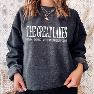 May include: A dark charcoal gray crewneck sweatshirt featuring the white text THE GREAT LAKES printed in a bold font, with the names HURON | ONTARIO | MICHIGAN | ERIE | SUPERIOR listed in a smaller white font directly underneath the main title.