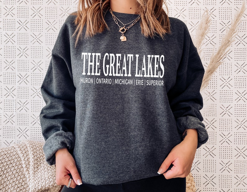May include: A dark charcoal gray crewneck sweatshirt featuring the white text THE GREAT LAKES printed in a bold font, with the names HURON | ONTARIO | MICHIGAN | ERIE | SUPERIOR listed in a smaller white font directly underneath the main title.