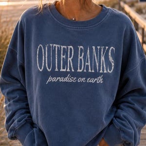 May include: A blue sweatshirt with the words "OUTER BANKS" in a white, distressed font. Below, the text reads "paradise on earth" in a cursive font. The sweatshirt has long sleeves and a crew neck.