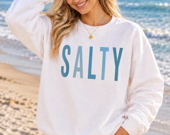 Könnte beinhalten: Ein weißes langärmeliges Sweatshirt mit Rundhalsausschnitt und dem Wort SALTY in großen, fetten, hellblauen Großbuchstaben auf der Brust. Das Kleidungsstück hat einen gerippten Kragen, Bündchen und Saum und wird mit hellblauen Jeansshorts kombiniert.