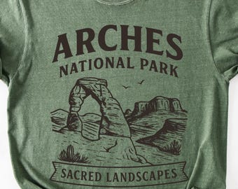 May include: A sage green short-sleeve t-shirt featuring a dark brown graphic print. The design displays the text ARCHES NATIONAL PARK above an illustration of a natural rock arch and desert landscape, with the text SACRED LANDSCAPES below.