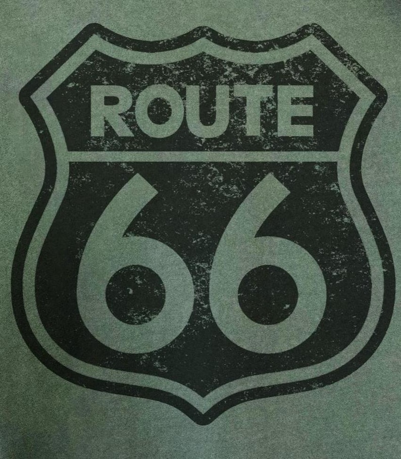 Puede incluir: Una superficie de tela verde oliva oscuro con un gr&aacute;fico negro de estilo desgastado del ic&oacute;nico emblema de escudo de la Ruta 66. El dise&ntilde;o incluye la palabra ROUTE en letras may&uacute;sculas negritas sobre el n&uacute;mero 66, todo dentro de un borde de escudo.