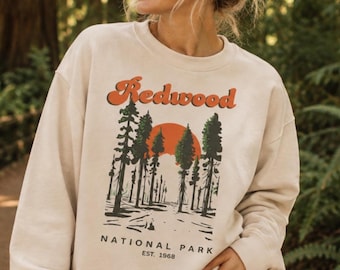 May include: A beige crewneck sweatshirt featuring a graphic print of a forest with tall trees and a large orange sun. The text "Redwood" appears in orange above the trees, with "NATIONAL PARK" and "EST. 1968" printed in black below the forest scene.