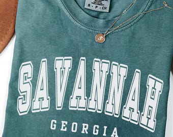 Puede incluir: Una camiseta Comfort Colors verde azulado con la palabra "SAVANNAH" en letras blancas y "GEORGIA" debajo. Un collar dorado con un pequeño colgante descansa sobre la camisa. La camisa es talla S-P-CH.