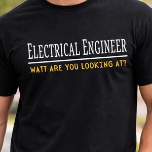 Puede incluir: Una camiseta negra de manga corta con el texto blanco "ELECTRICAL ENGINEER" sobre una línea horizontal blanca, con el texto amarillo "WATT ARE YOU LOOKING AT?" impreso debajo. La camiseta tiene cuello redondo y un corte estándar.
