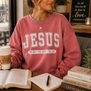 May include: A dusty rose crewneck sweatshirt featuring the white text JESUS in a collegiate font, with the phrase THE WAY &bull; THE TRUTH &bull; THE LIFE printed in a white oval below. The garment has long sleeves and a ribbed collar and cuffs.