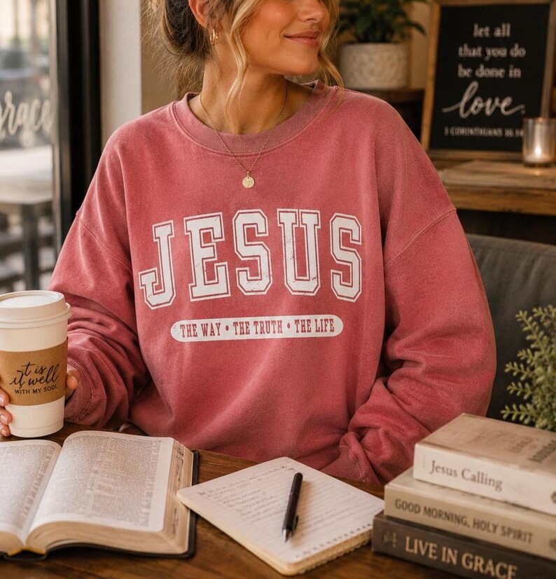 May include: A dusty rose crewneck sweatshirt featuring the white text JESUS in a collegiate font, with the phrase THE WAY &bull; THE TRUTH &bull; THE LIFE printed in a white oval below. The garment has long sleeves and a ribbed collar and cuffs.