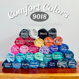 May include: A pyramid stack of rolled Comfort Colors 9018 Youth Midweight T-Shirts in various colors including gray, white, black, pepper, watermelon, crimson, butter, melon, terracotta, neon pink, blossom, crunchberry, orchid, violet, flo blue, chambray, lagoon blue, chalky mint, island reef, seafoam, island green, topaz blue, china blue, washed denim, denim, royal caribbean, ice blue, true navy, granite, and blue jean.