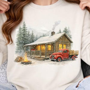 Pode incluir: Camisola creme com uma cena de inverno. Uma cabana de madeira aconchegante com janelas iluminadas e uma picape vermelha são retratadas em uma floresta nevada. Uma fogueira queima em frente à cabana, criando uma atmosfera acolhedora.