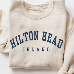 May include: A folded, beige sweatshirt with the words "HILTON HEAD ISLAND" printed in navy blue arched across the front. The sweatshirt has a crew neck and long sleeves.