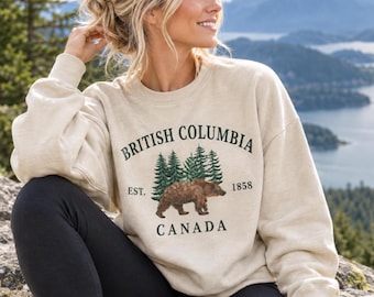 Pode incluir: Uma camisola bege de gola redonda com um gráfico de um urso castanho a caminhar em frente a pinheiros verdes. O texto diz British Columbia, Est. 1858 e Canada em verde escuro. A camisola tem mangas compridas e uma gola canelada.